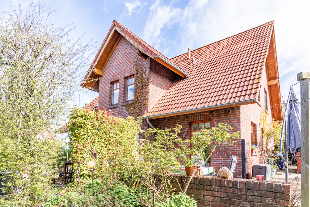 A two-story brick house with a red tile roof, surrounded by lush greenery and a brick wall.