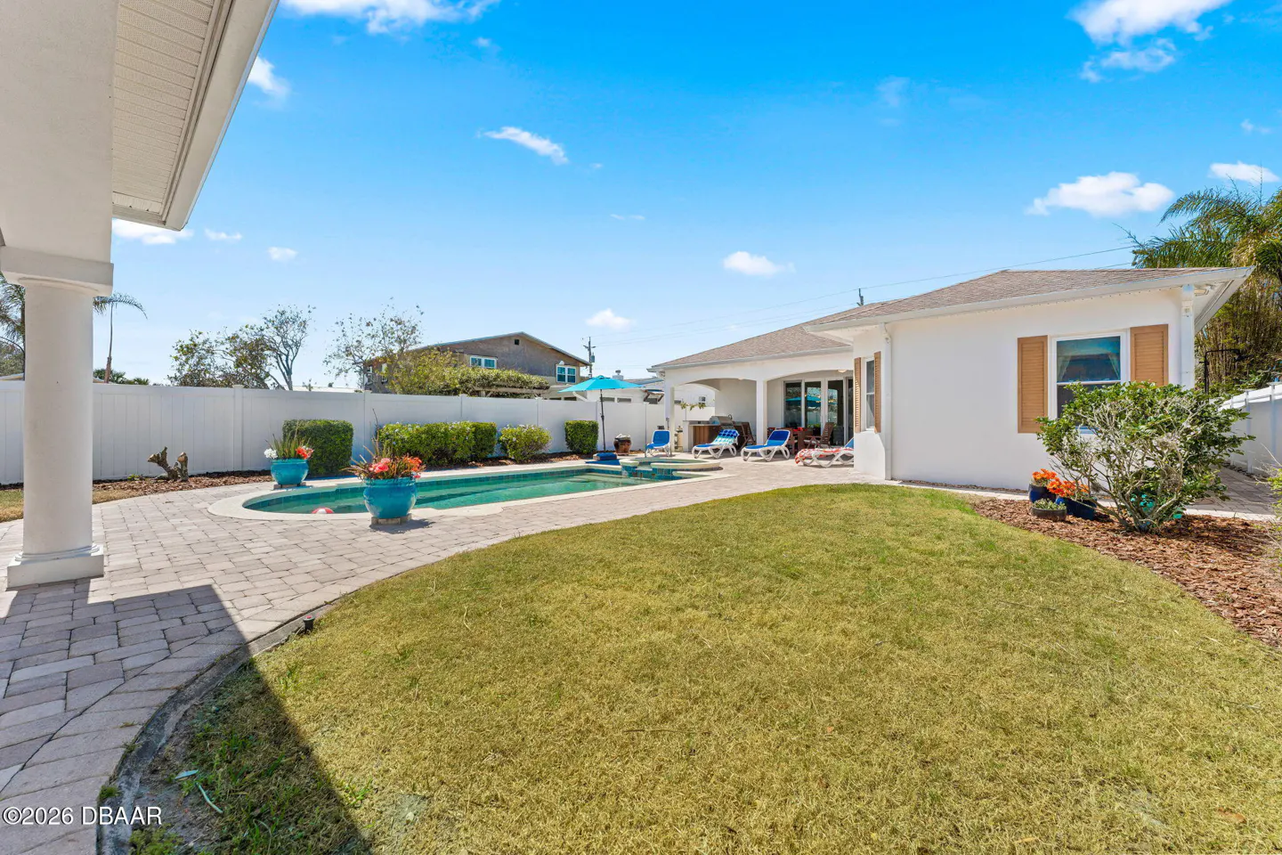 A backyard with a pool, lawn, and white house under a blue sky. Lounge chairs are near the pool.