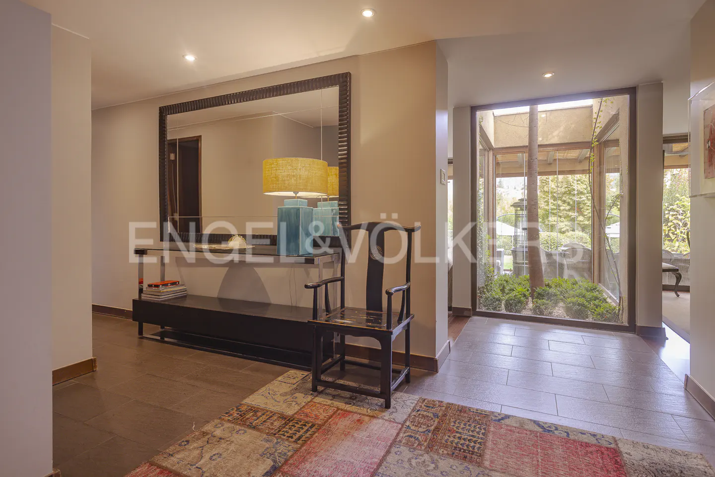 A foyer with a large mirror, a black console table with a blue lamp, and a wooden chair on a colorful rug. A window shows a garden.