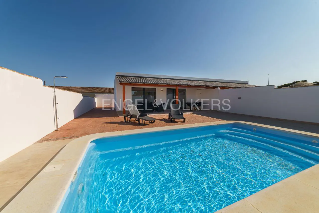 A backyard with a blue swimming pool, two lounge chairs, and a white house under a clear blue sky.