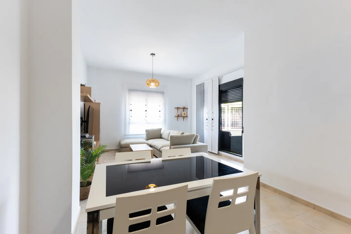 A bright, modern living and dining area with a black-topped table and white chairs in the foreground. A beige sectional sofa and TV stand are visible in the background.