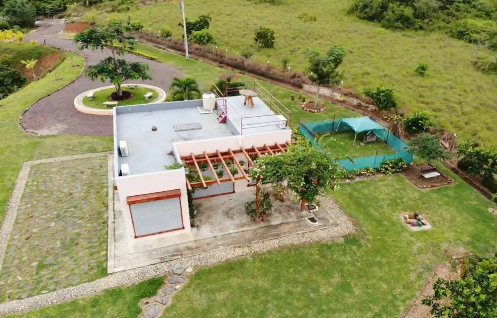 Aerial view of a modern, single-story house with a flat roof, surrounded by green grass and trees. A pergola covers the patio.