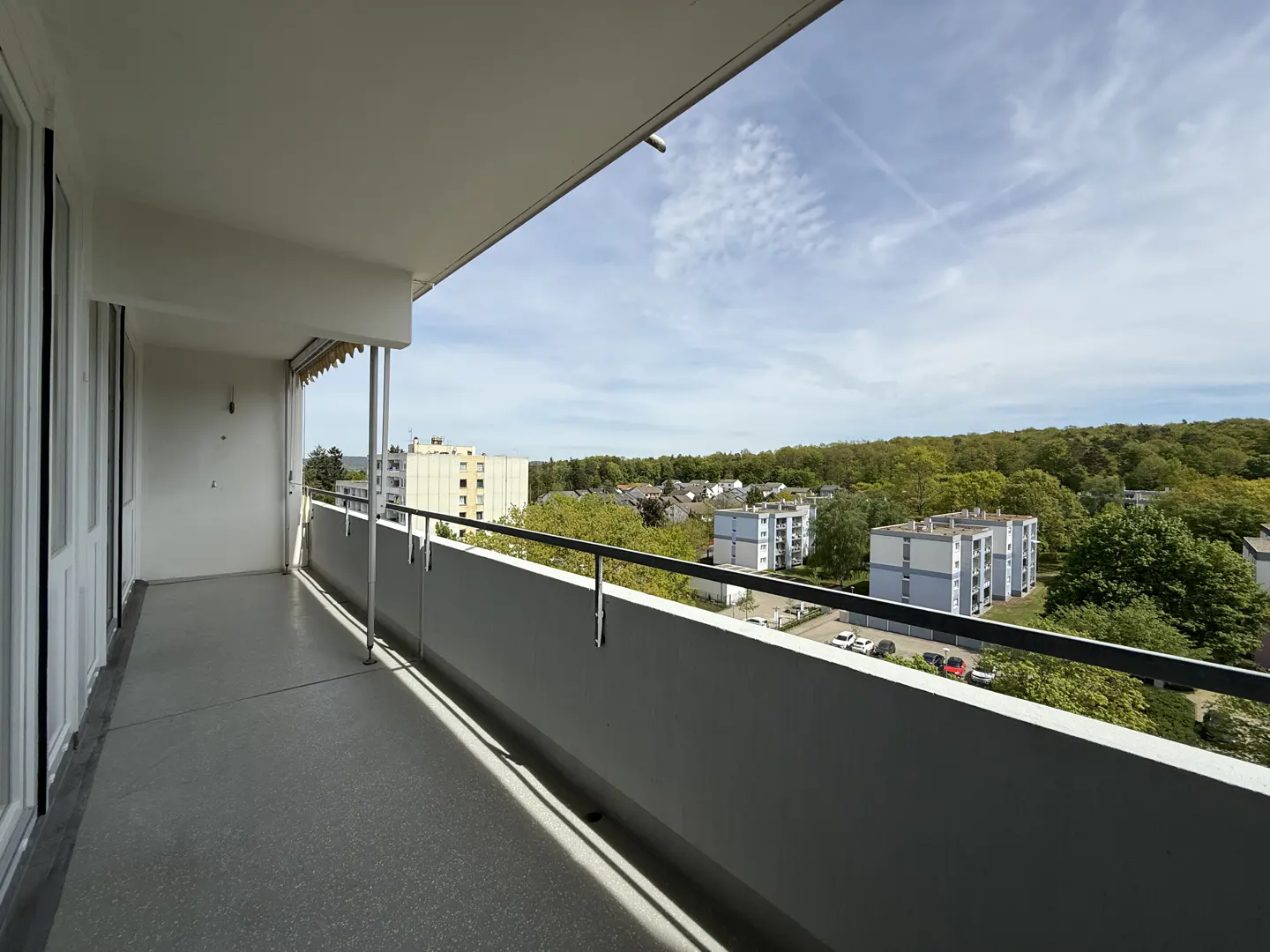Spacious apartment balcony with a grey floor, metal railing, and a scenic view of residential buildings and green trees.