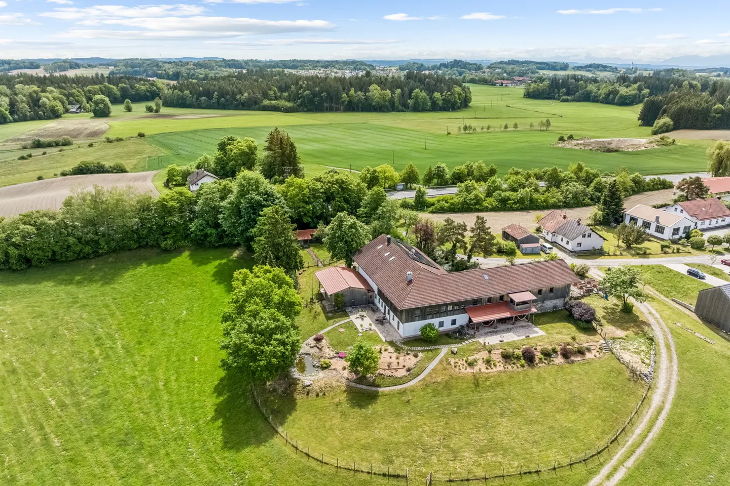 Aerial view of a large farmhouse with a brown roof, surrounded by lush green fields, trees, and a gravel driveway.