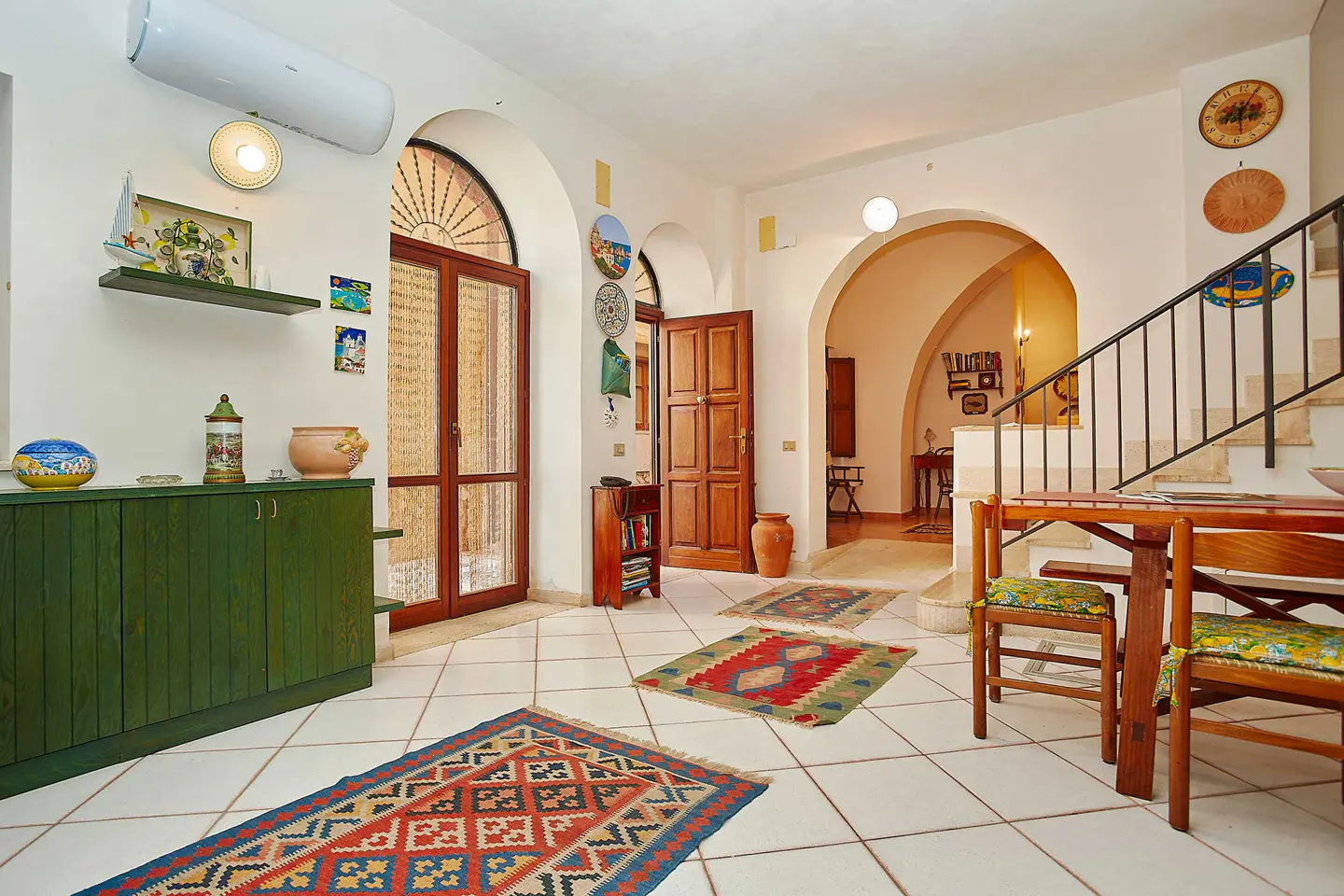 Bright, airy room with white tile floors, arched doorways, and colorful rugs. Green cabinet, wooden table and chairs, and staircase visible.