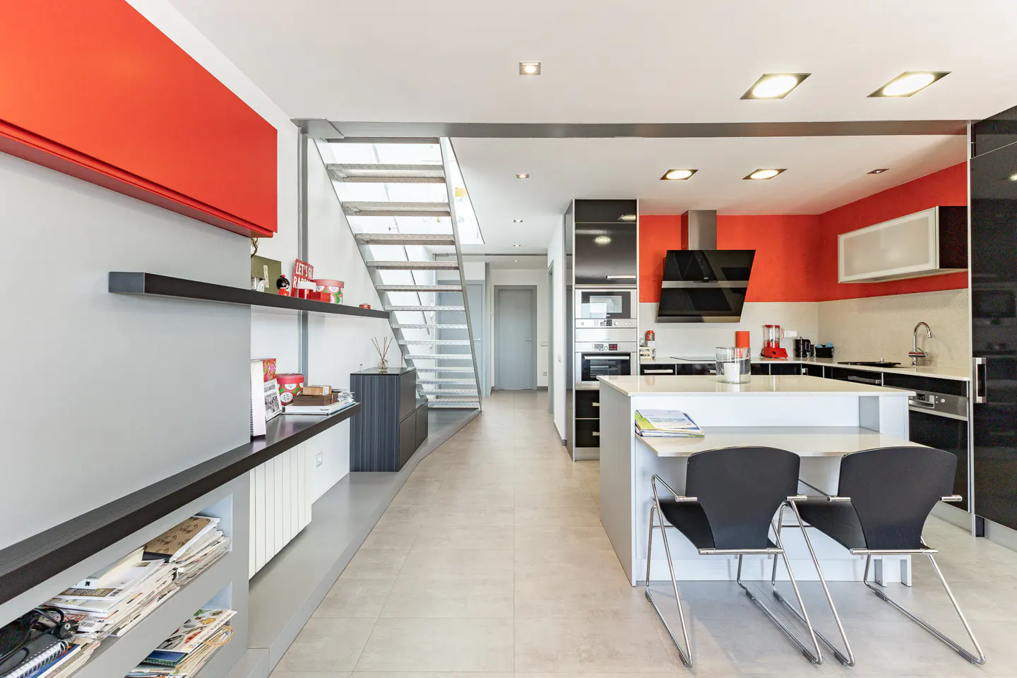 A modern kitchen with a white island, black chairs, and stainless steel appliances. Red accents on the walls and a metal staircase are visible.