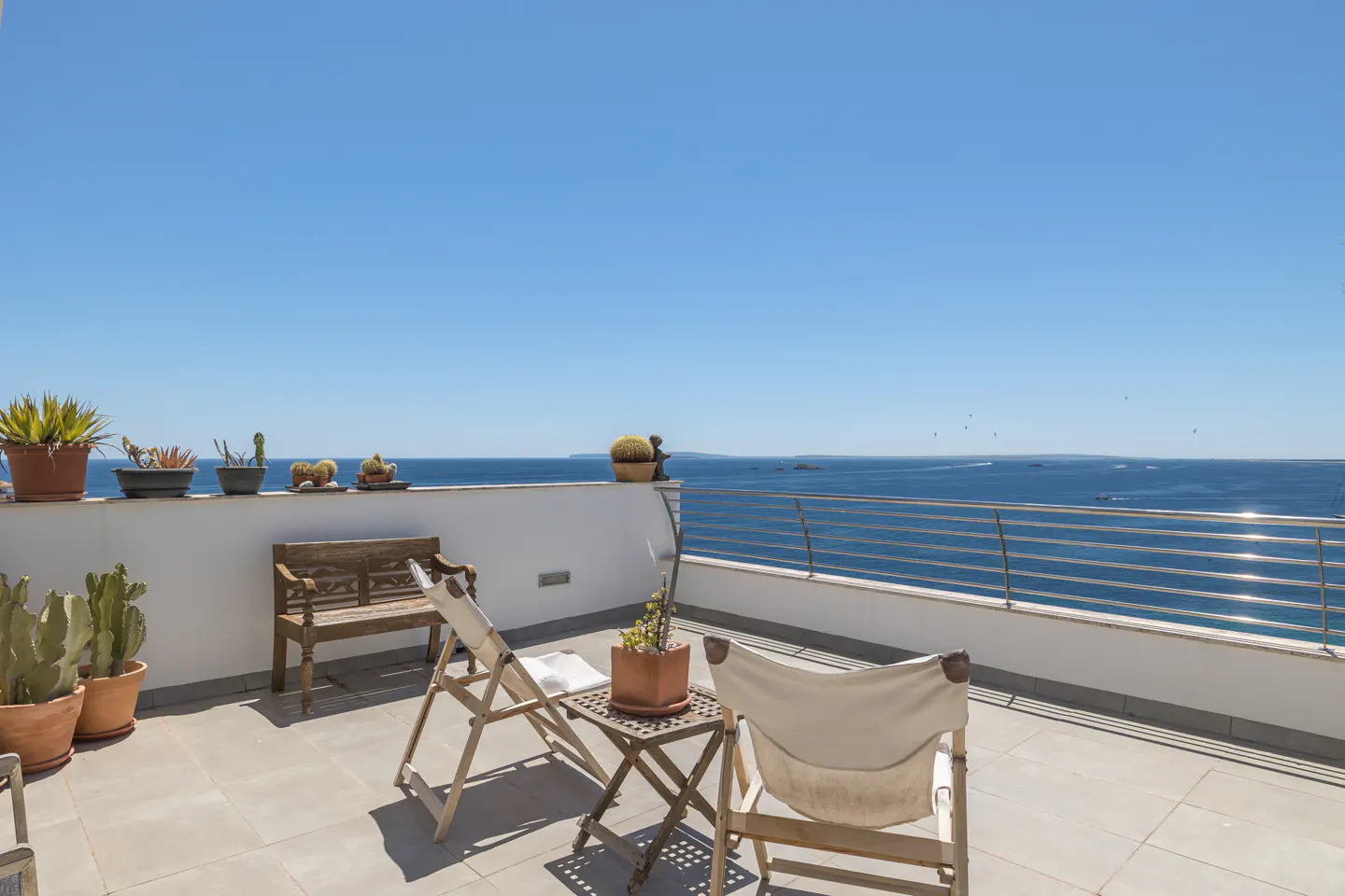 Sunny terrace with a wooden bench, two folding chairs, potted cacti, and a metal railing overlooking the blue ocean.