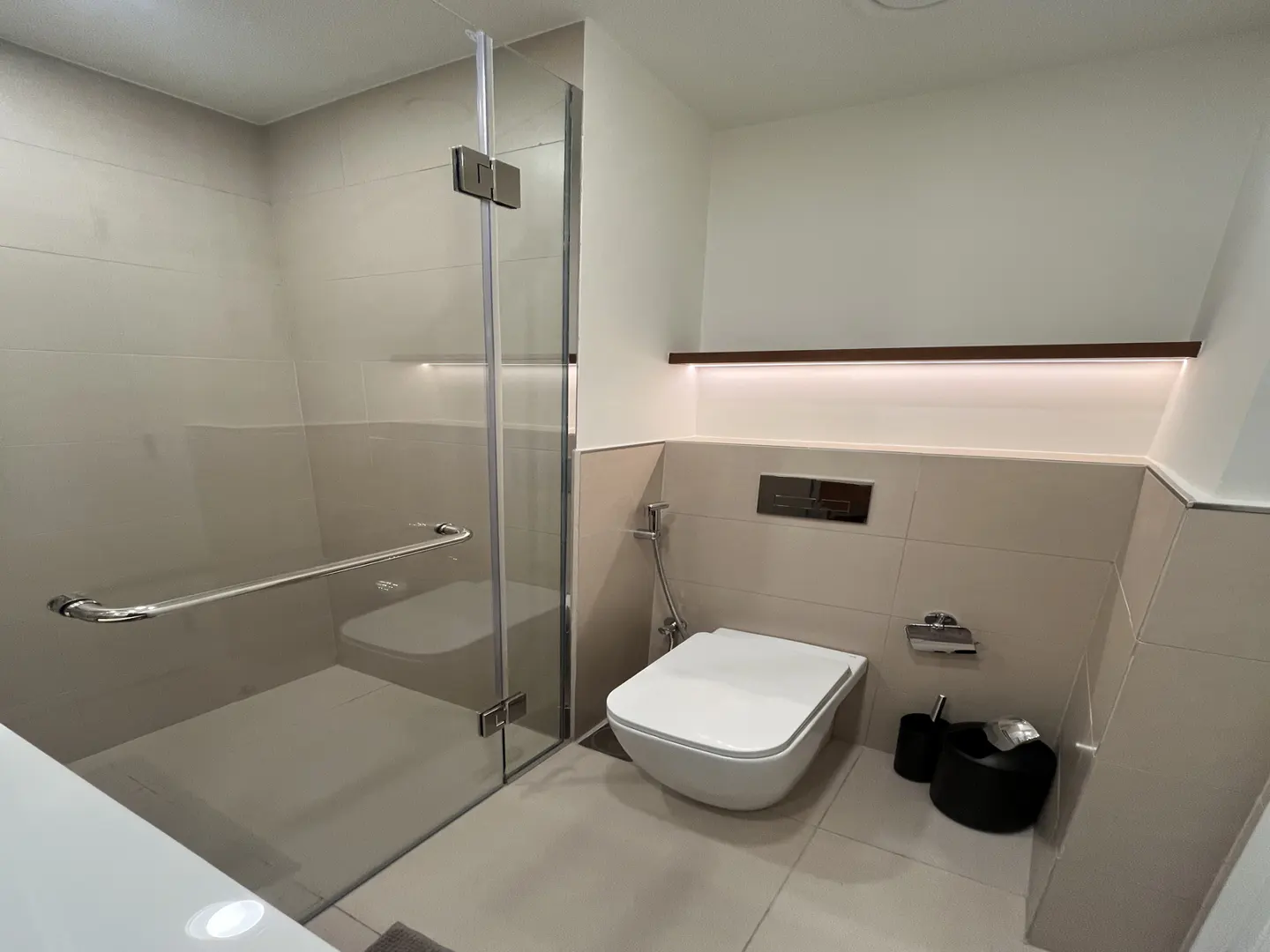 Modern bathroom with a glass shower, white toilet, and beige tile walls. A dark wood shelf with lighting is above the toilet.