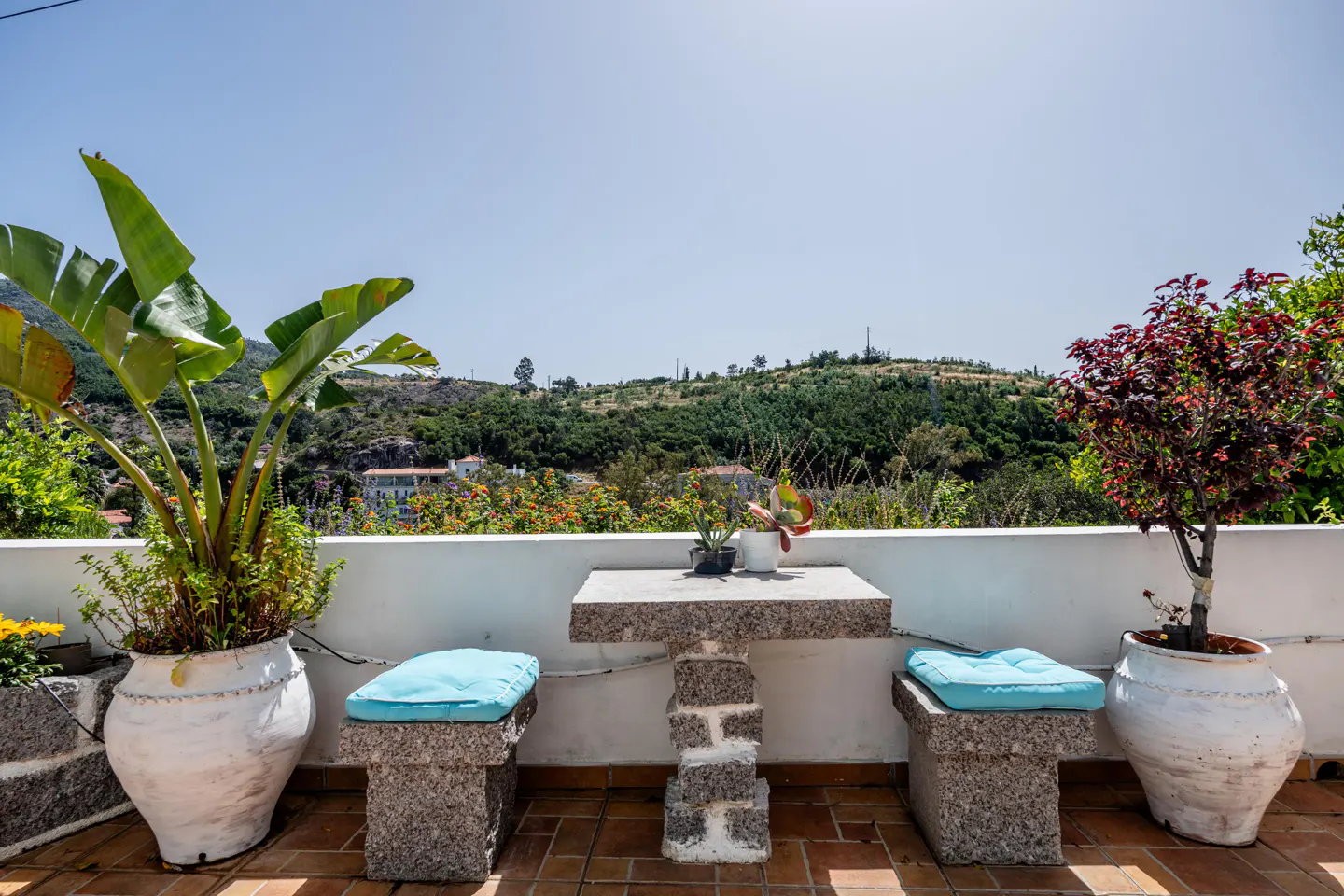 Outdoor patio with stone table and benches with blue cushions. Potted plants frame a view of green hills under a clear blue sky.