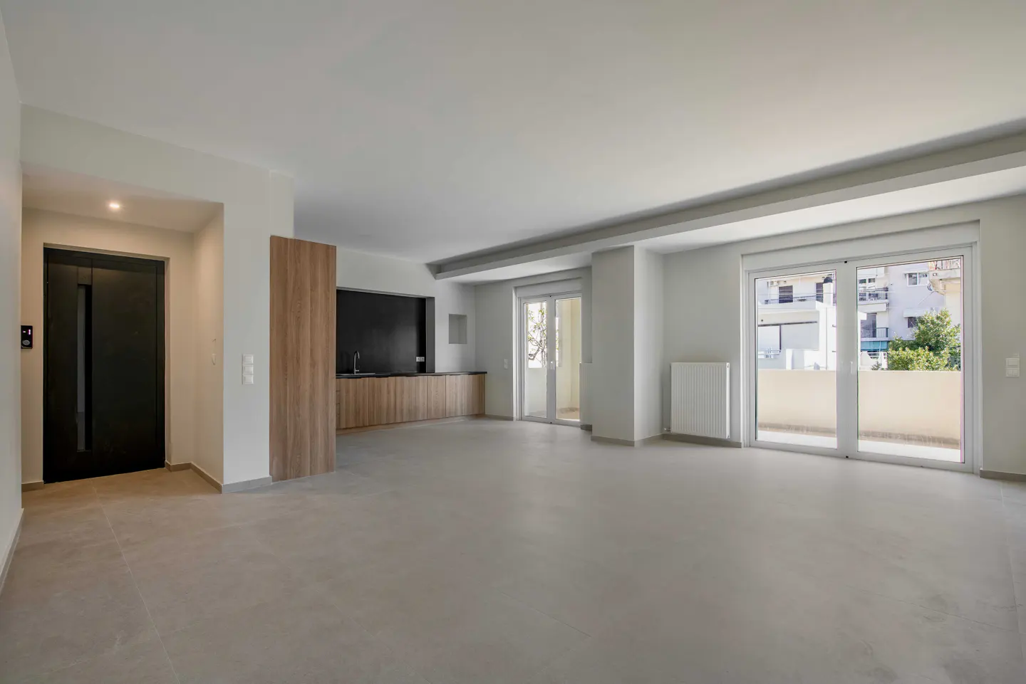 Bright, empty apartment with gray floors, white walls, and a black accent wall in the kitchen area. Balcony doors let in natural light.