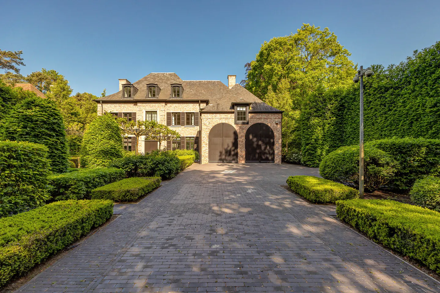 A large, light-brick house with black shutters and a gray roof, surrounded by green trees and manicured hedges. A brick driveway leads to the house.