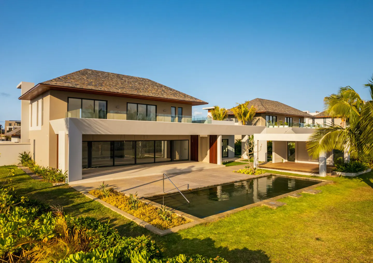 Exterior view of a modern, two-story home with a pool, lawn, and blue sky. The house is beige with a brown roof and large windows.
