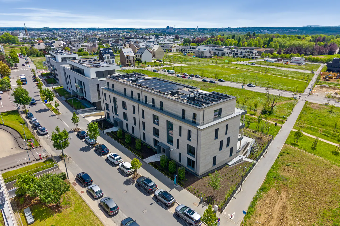 Aerial view of modern, light-stone apartments with solar panels, parked cars, and green spaces under a blue sky.