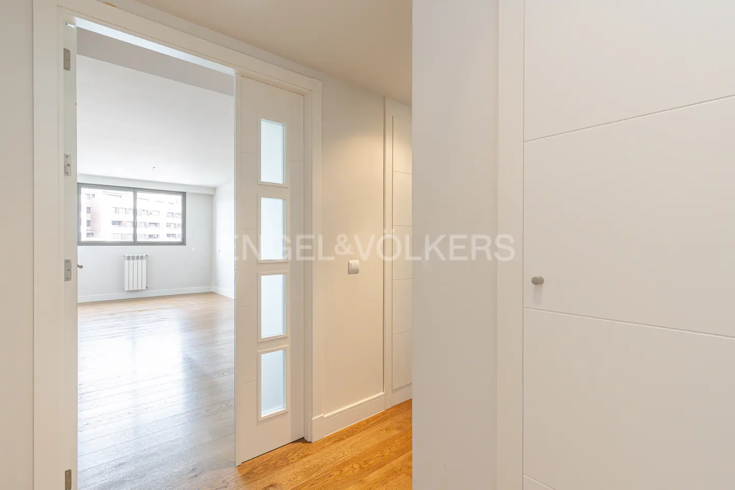Interior view of a bright hallway with wood floors, white walls, and doors. A room with a window is visible through an open doorway.
