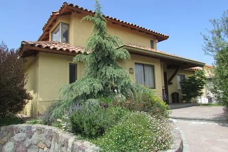 Two-story tan house with a red tile roof, a stone wall, and a large evergreen tree in the front yard.
