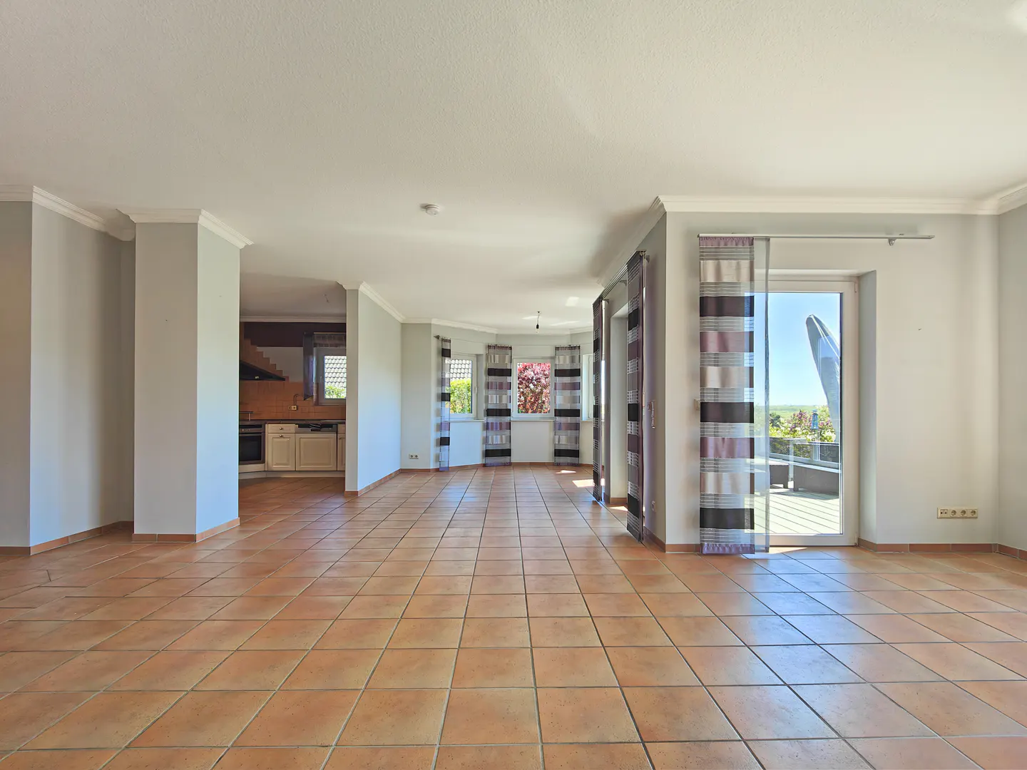 Empty room with terracotta tile floor, gray walls, white trim, and striped curtains. A kitchen is visible in the background. Balcony access is on the right.