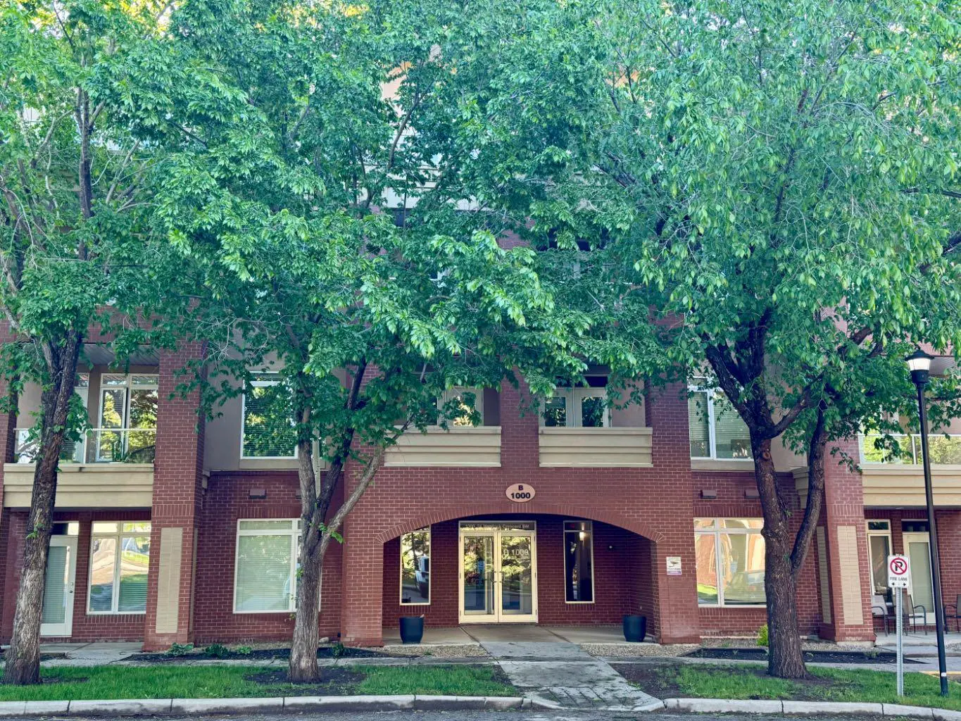 A three-story red brick apartment building with a central arched entrance, framed by large, leafy green trees.