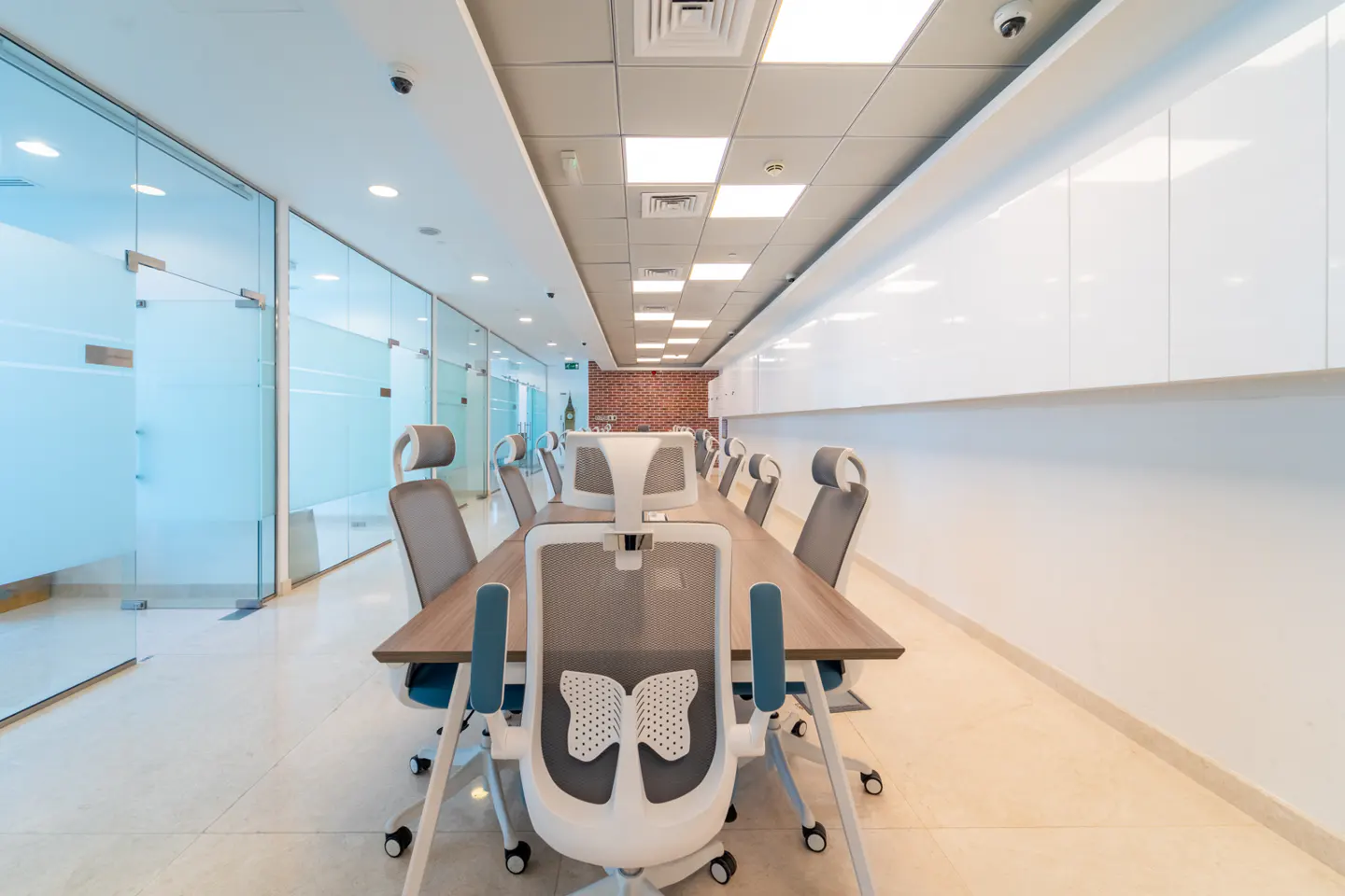 Bright, modern office with a long wooden conference table surrounded by ergonomic chairs. Glass-walled offices line one side.