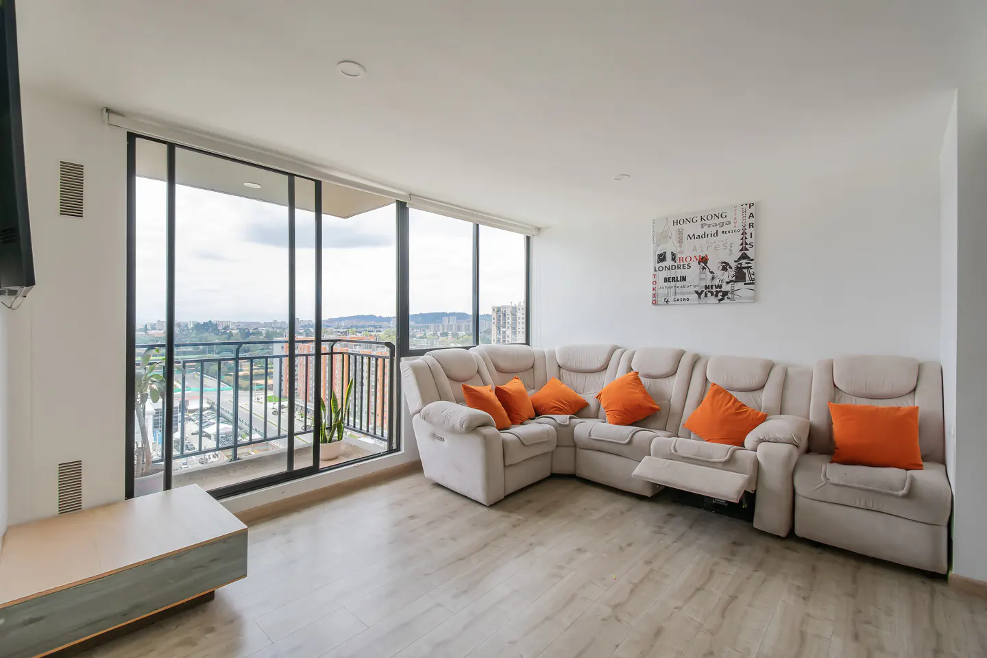Bright living room with beige sectional sofa, orange pillows, and city view from a balcony with black railings.