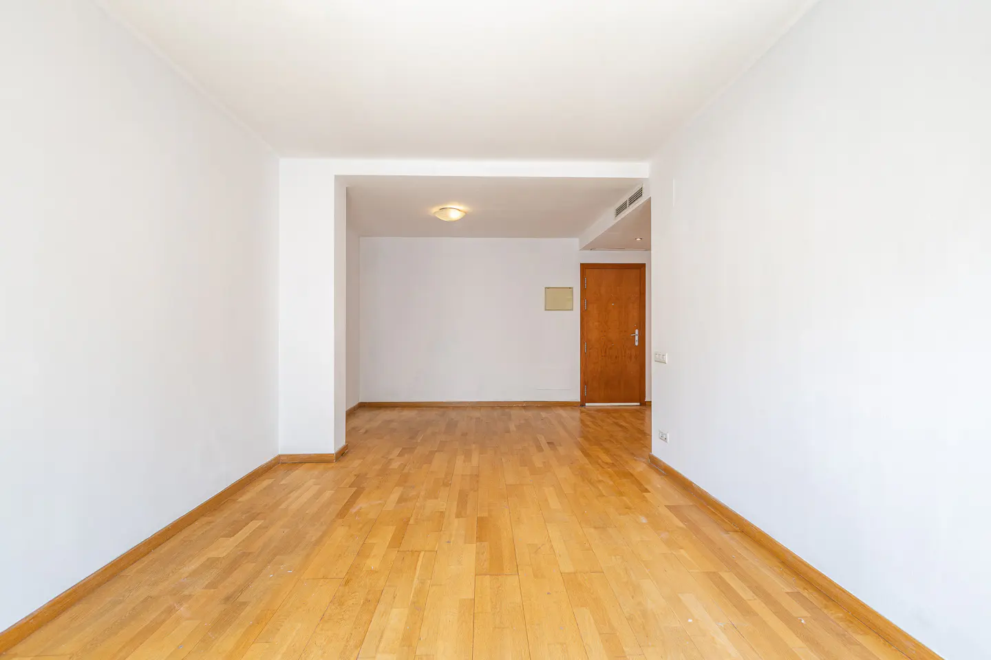 Empty room with white walls, light wood floors, and a doorway leading to another room with a brown door.
