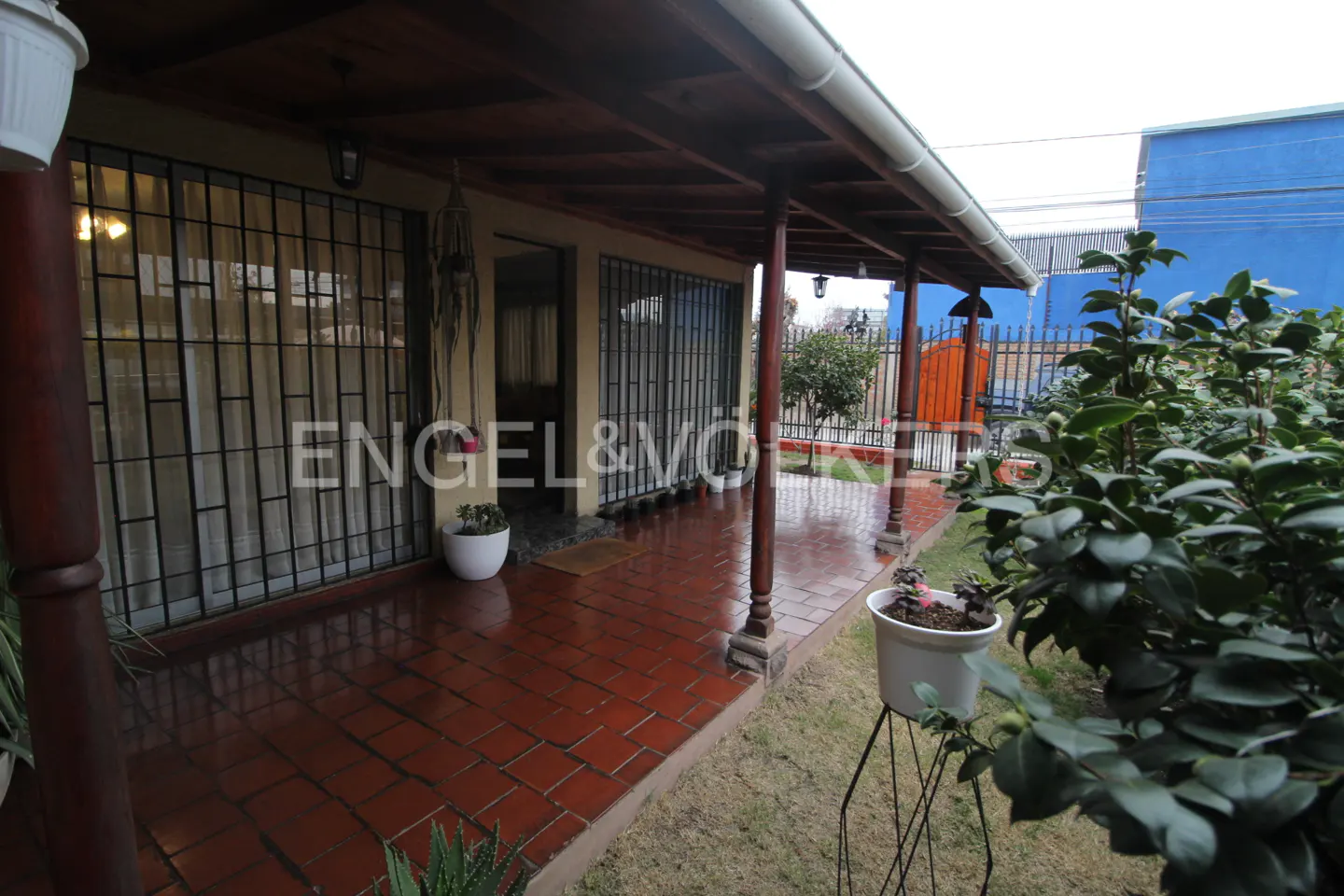 Covered patio with red tile flooring, wooden pillars, and large windows with security bars, overlooking a small garden.