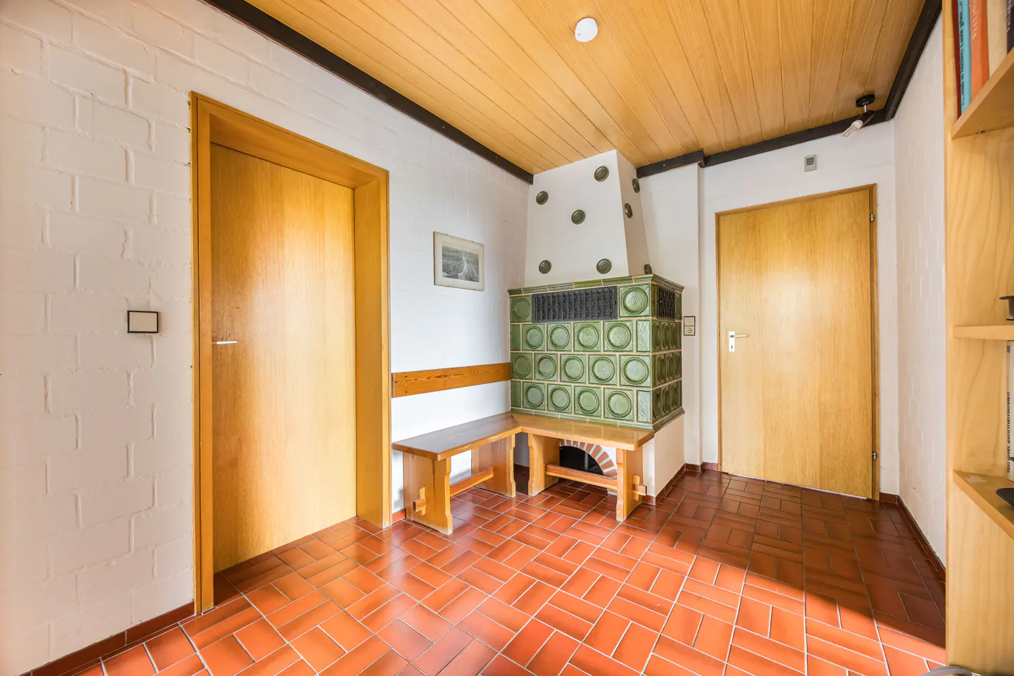 Hallway with terracotta tile floor, wood-paneled ceiling, and a green tiled stove with a bench. Two wooden doors are visible.