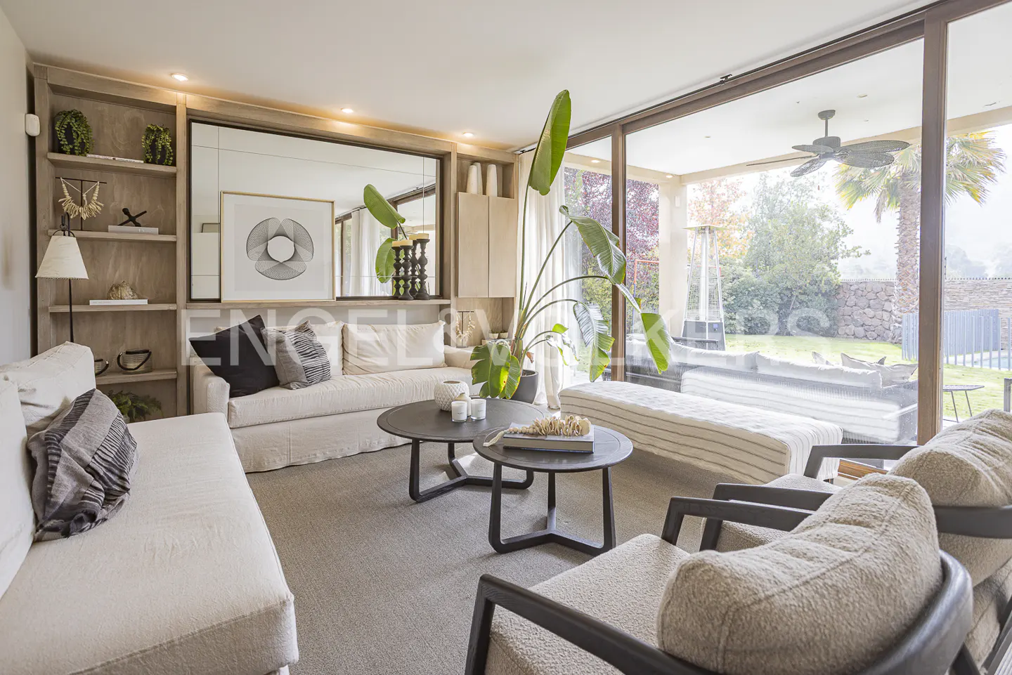 Living room with beige sofas, dark wood tables, and a large window overlooking a patio and garden.
