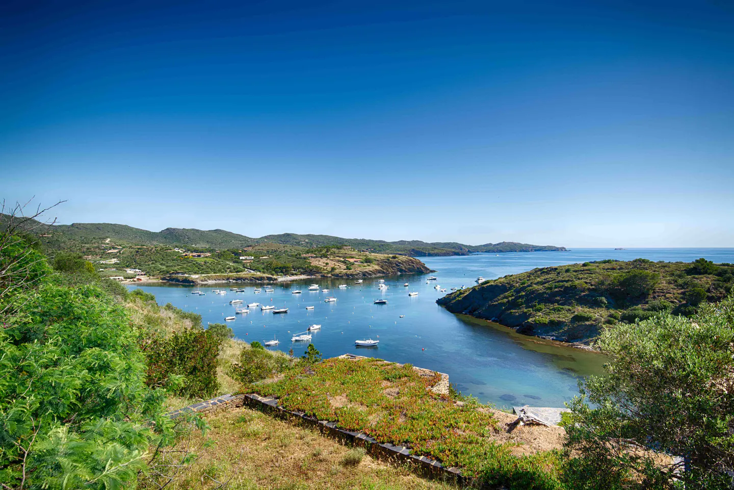 A scenic view of a calm blue bay filled with small white boats, surrounded by green hills under a clear blue sky.