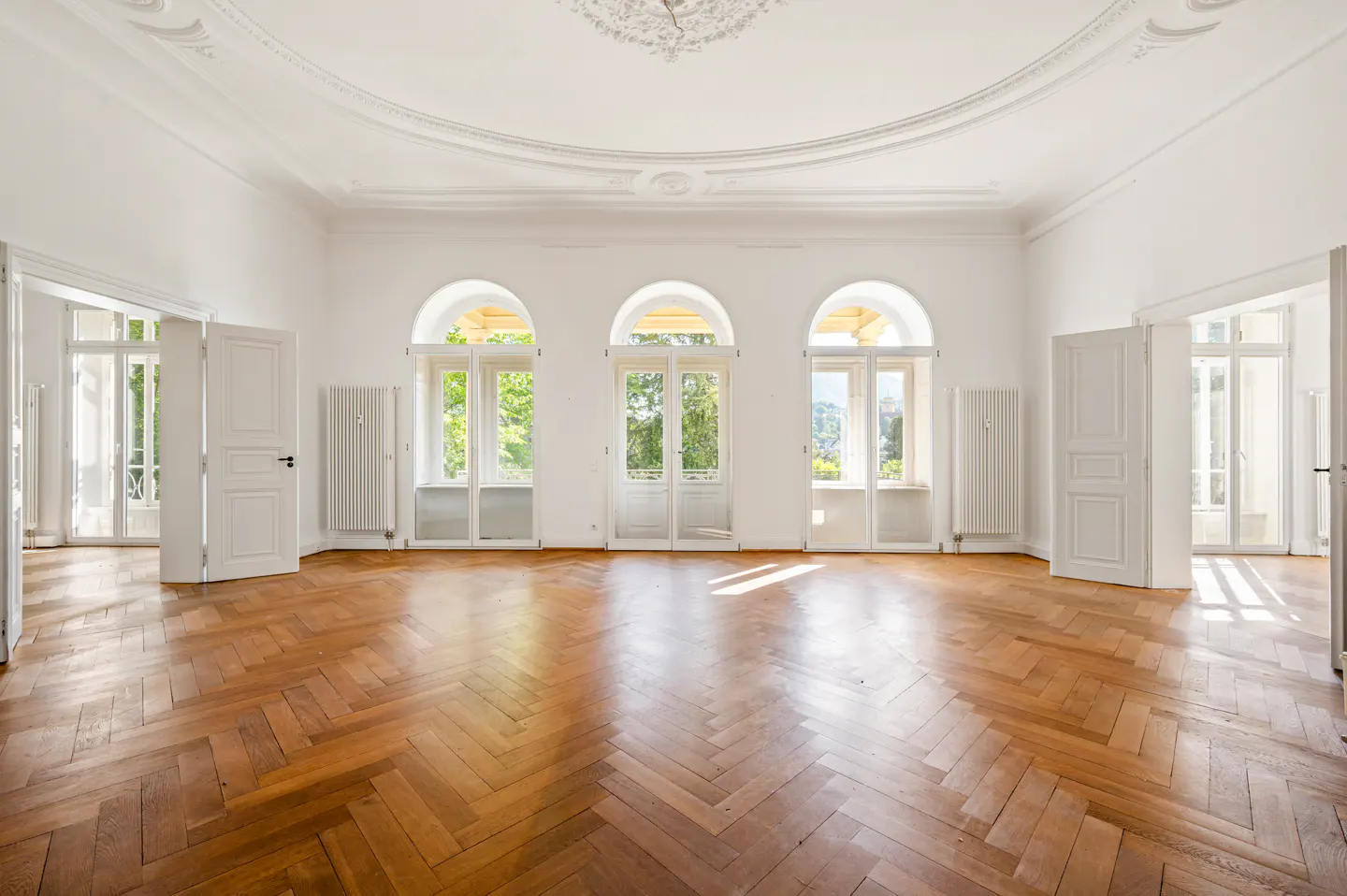Bright, empty room with herringbone wood floors, white walls, and three arched windows overlooking greenery. White doors and radiators are visible.