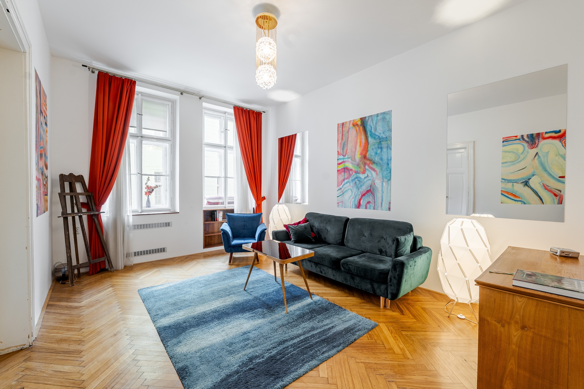 Bright living room with a dark green sofa, blue armchair, blue rug, wooden floors, and vibrant abstract art on white walls. [AI-generated text]