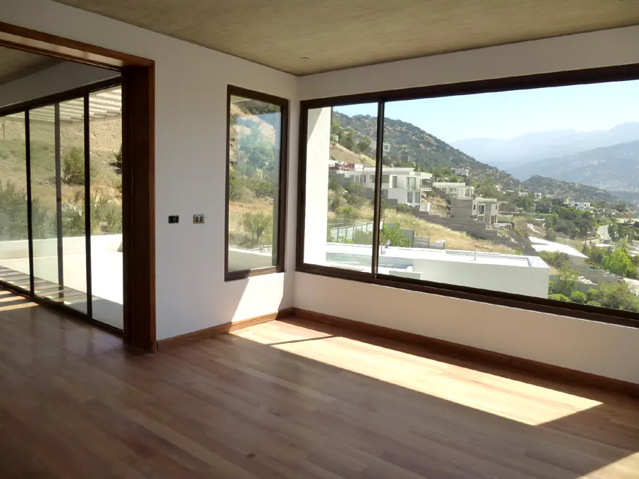 An empty room with light wood floors and large windows. Outside, a hillside with houses is visible.