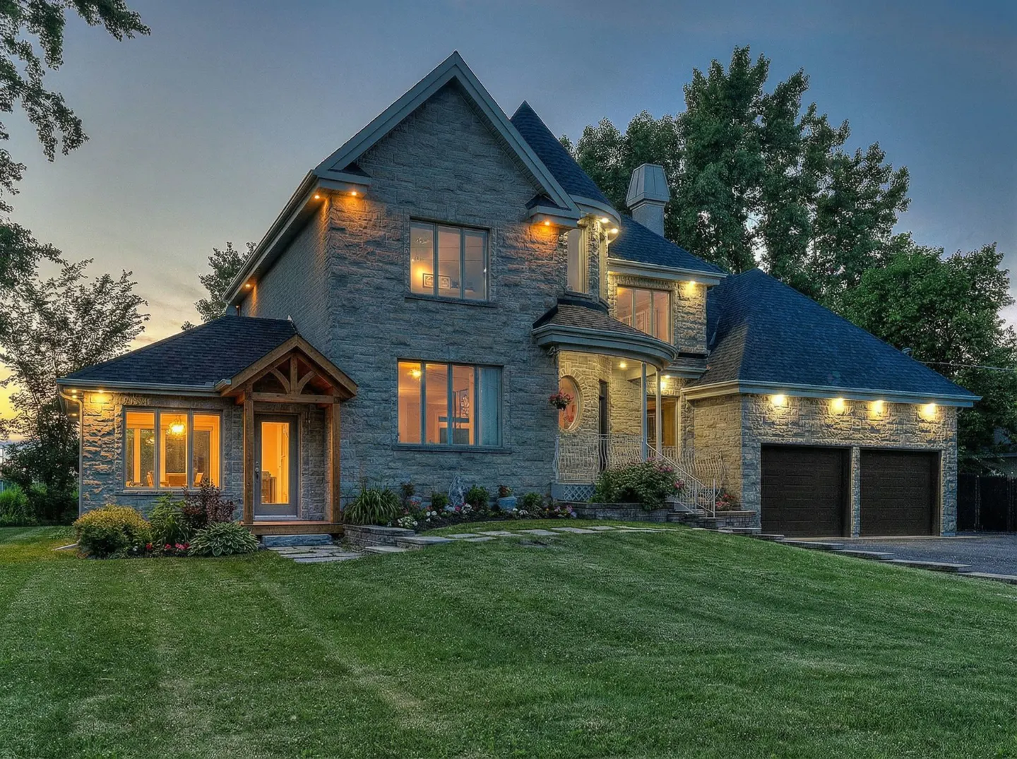 Two-story stone house with a dark roof, a two-car garage, and a green lawn at dusk.