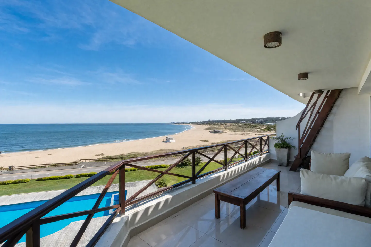 Balcony view of ocean, beach, and pool. Brown wood railing and bench. White sofa with pillows. Blue sky.