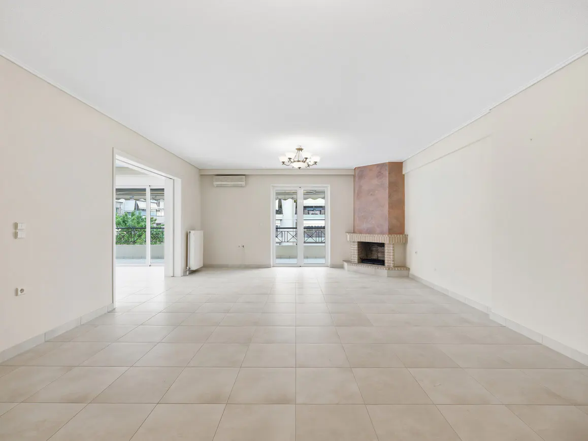 Empty living room with beige tile floor, white walls, fireplace, chandelier, and balcony access.