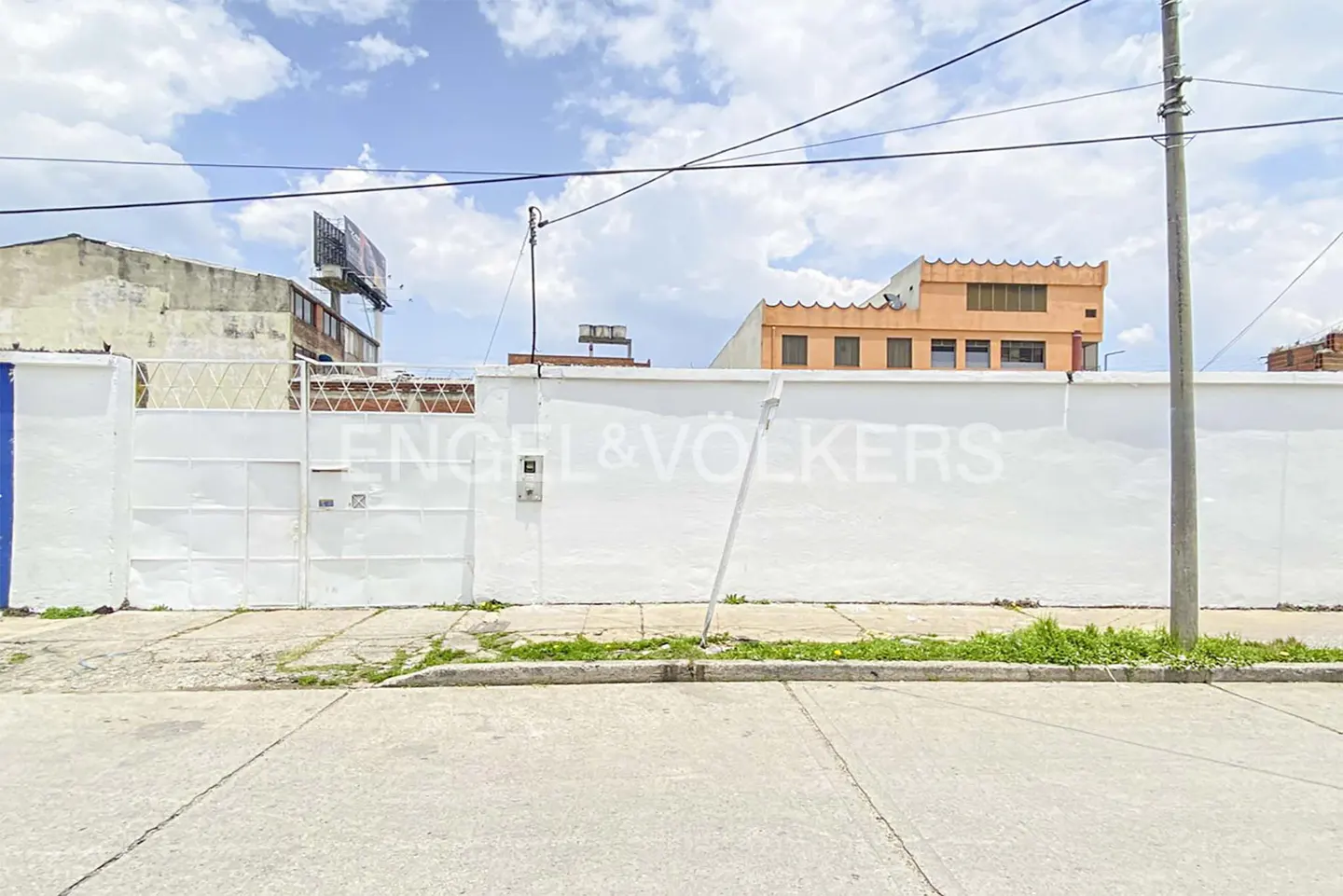 Real estate view of a white wall with Engel & Völkers logo, a gate, and buildings under a cloudy sky.