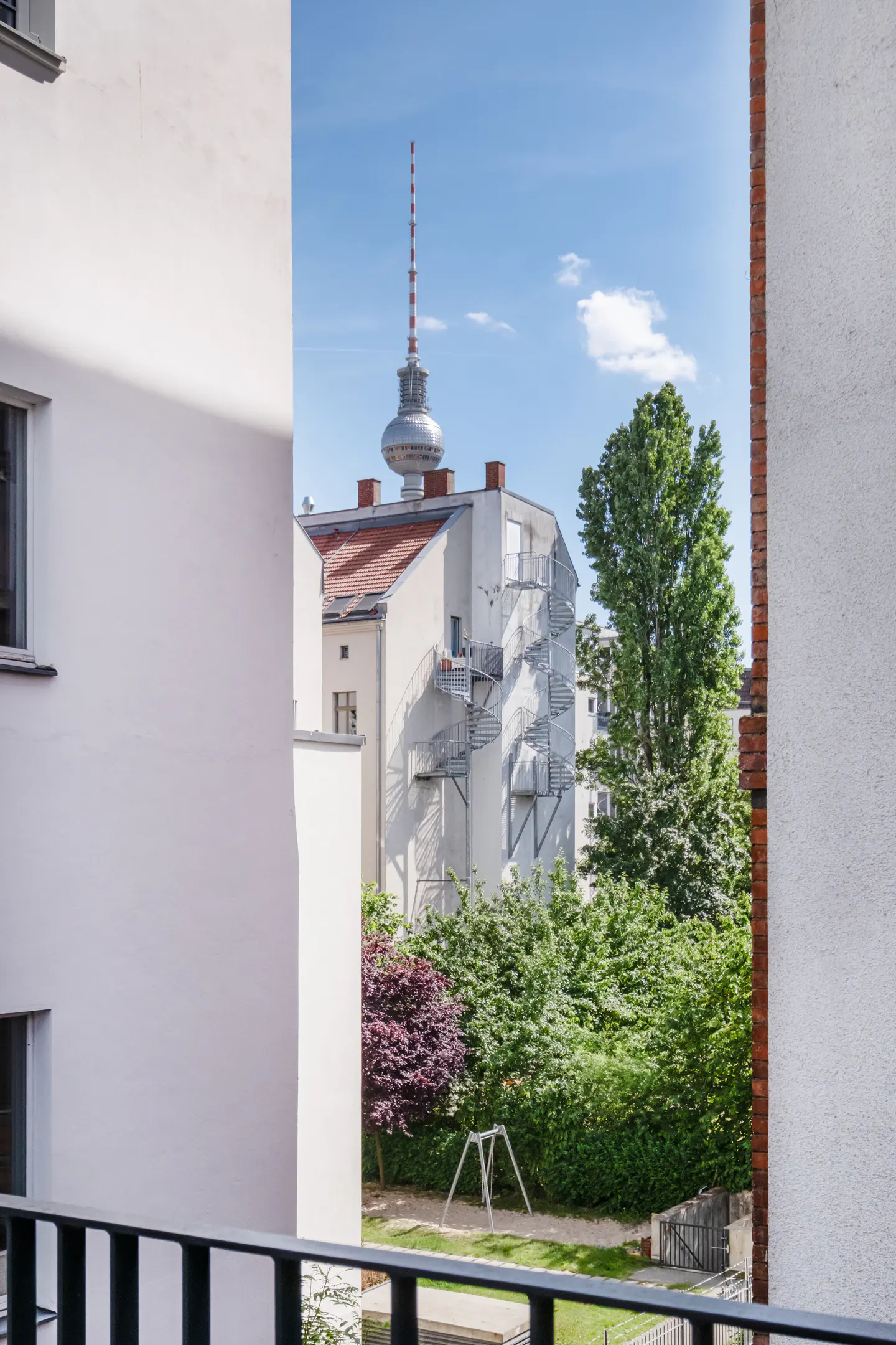 View from a balcony looking toward the Berlin TV Tower rising above residential buildings, trees, and a small playground.