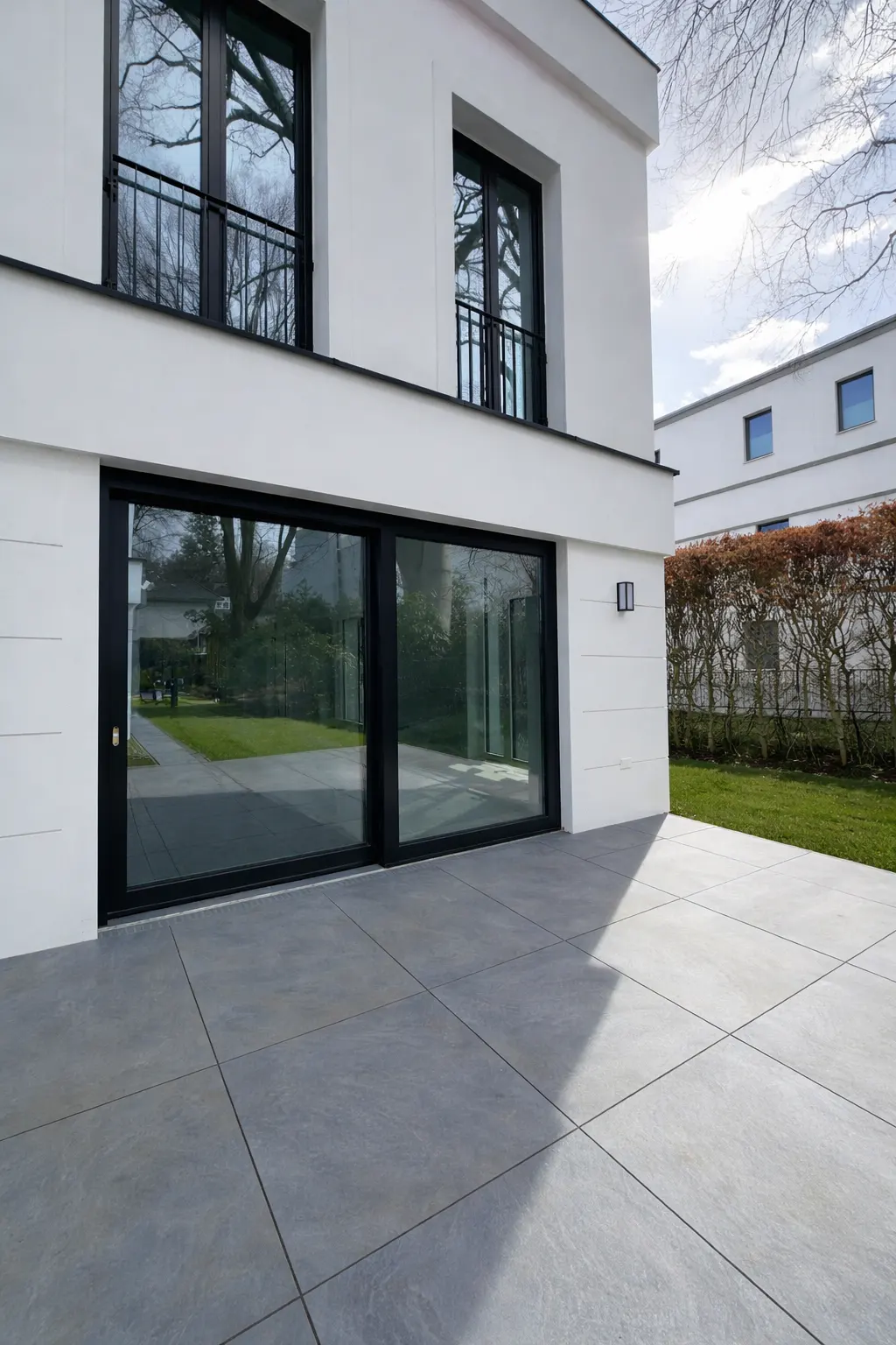 Modern white house with black framed windows and sliding glass doors. Gray tiled patio leads to a green lawn and manicured hedges.
