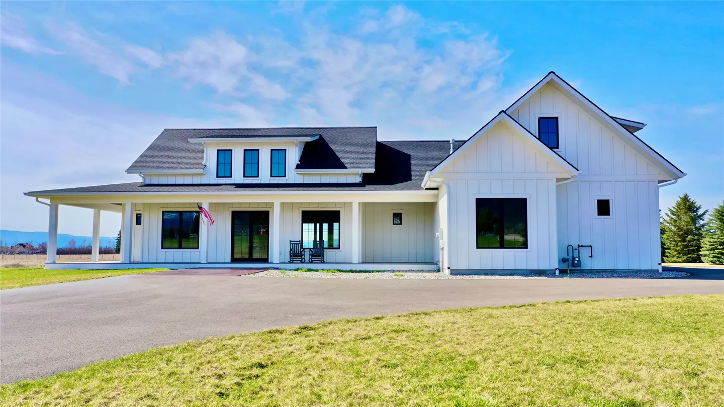 A modern farmhouse with white siding, a dark roof, and black-framed windows sits on a green lawn under a blue sky.