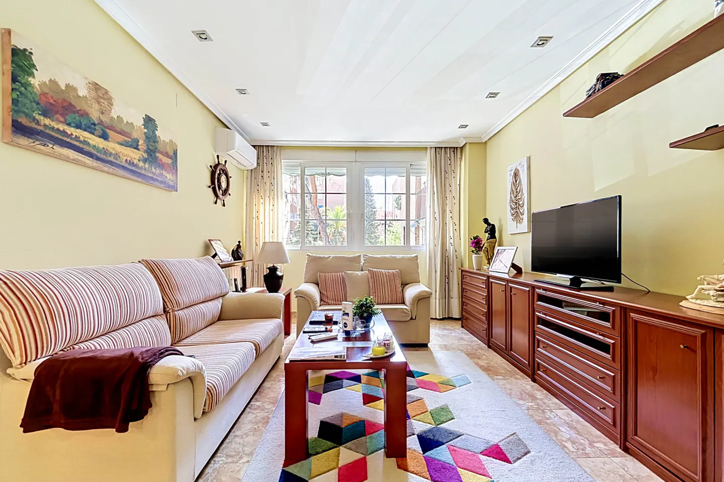 Living room with yellow walls, a striped sofa, a beige loveseat, and a colorful geometric rug. A TV sits on a wooden cabinet.