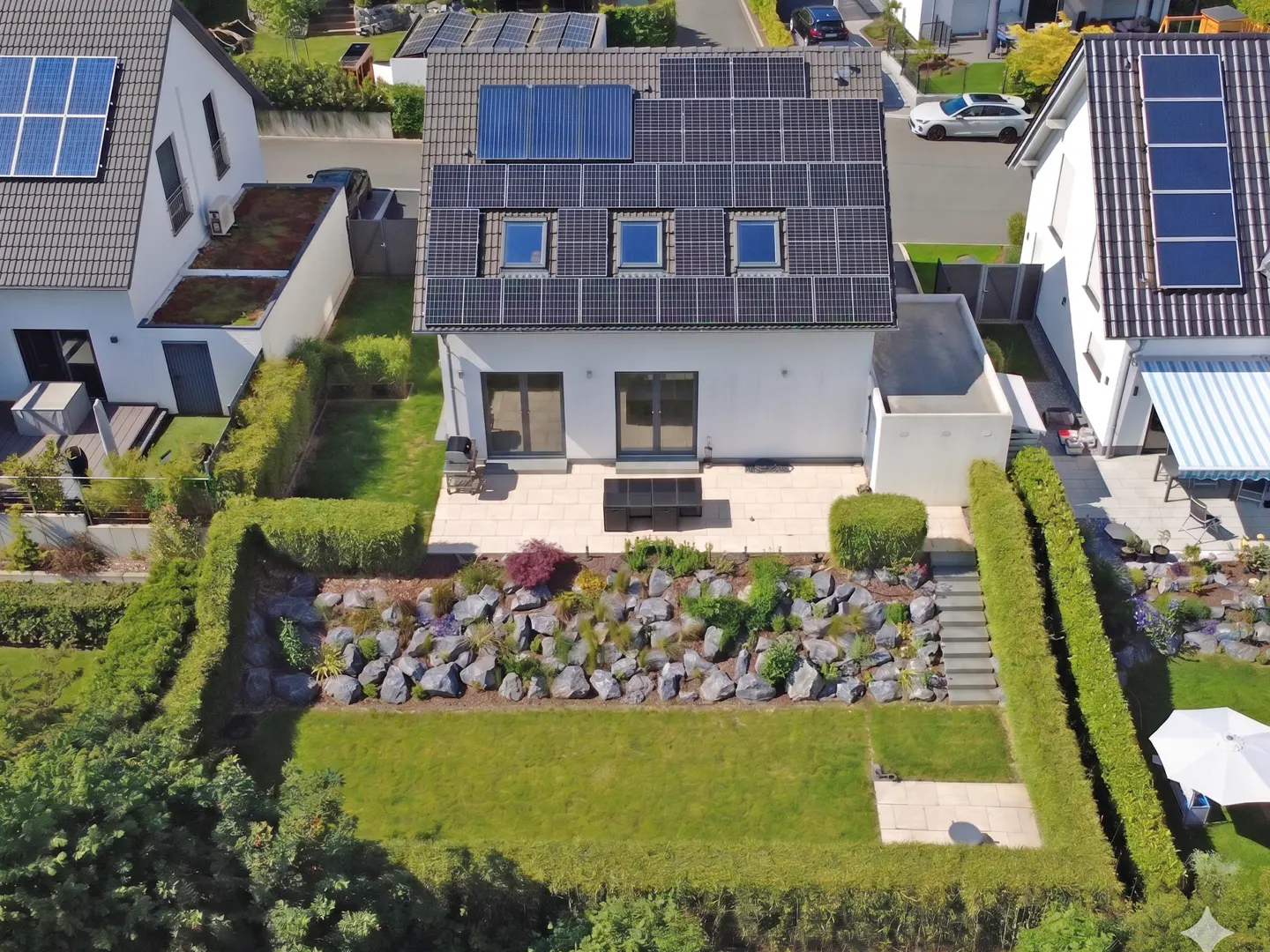 Aerial view of a modern white house with solar panels, a patio, and a landscaped garden with rocks and green hedges.