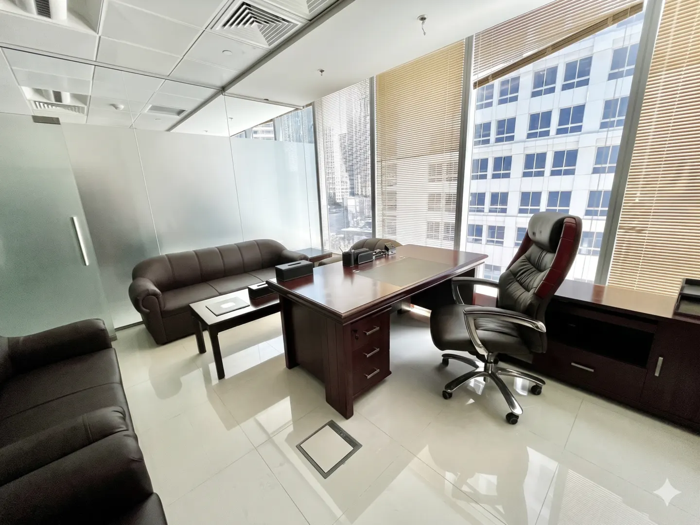 A modern office space with brown leather sofas, a large wooden desk, and a black executive chair. A large window shows a city view.