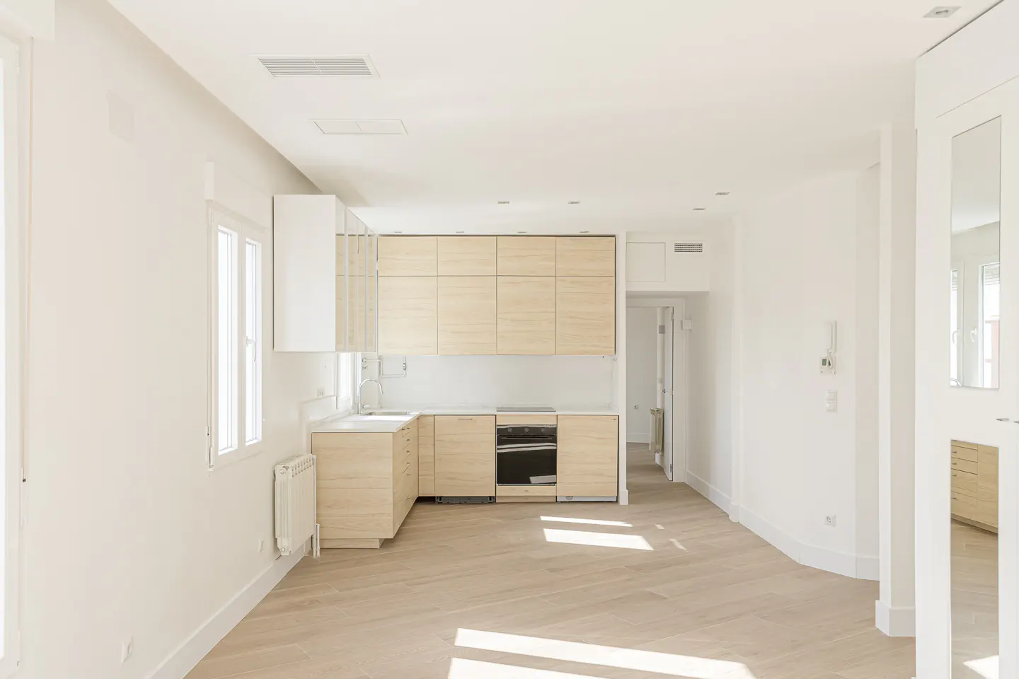 Bright, empty apartment with light wood floors and white walls. A modern kitchen with light wood cabinets is on the left. A doorway is visible in the background.