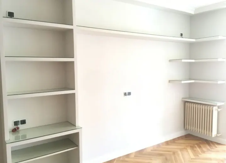 Bright room with white built-in shelves and a radiator. The shelves have glass tops. A herringbone wood floor adds warmth.