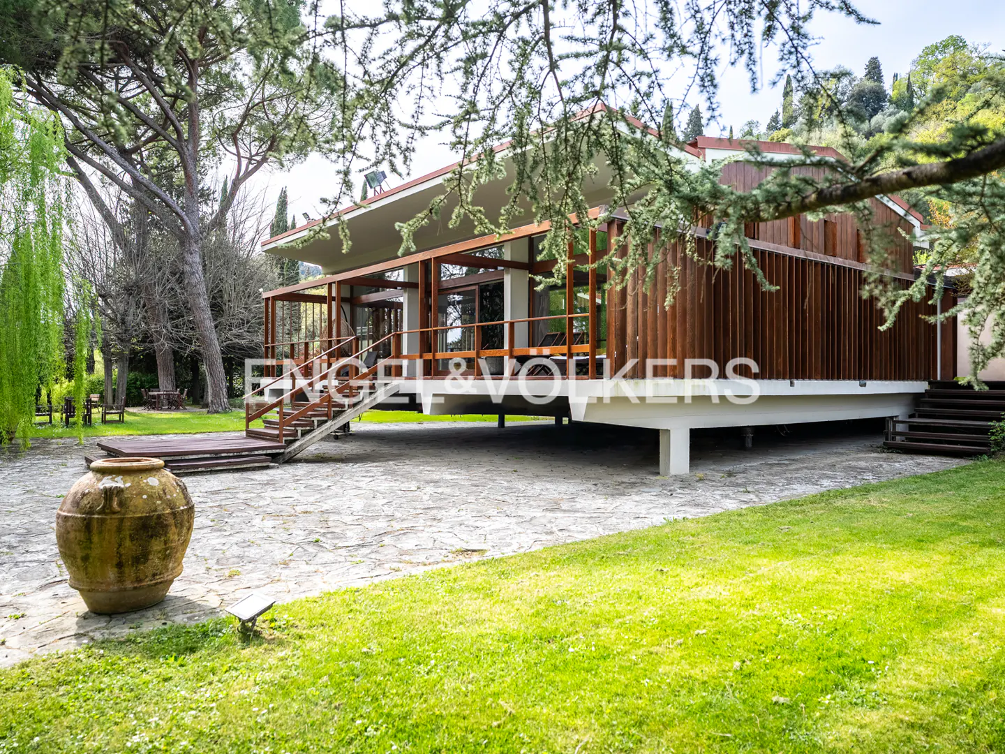 Exterior view of a modern, elevated wood and glass house with a stone patio and green lawn. A large, weathered pot sits in the foreground.