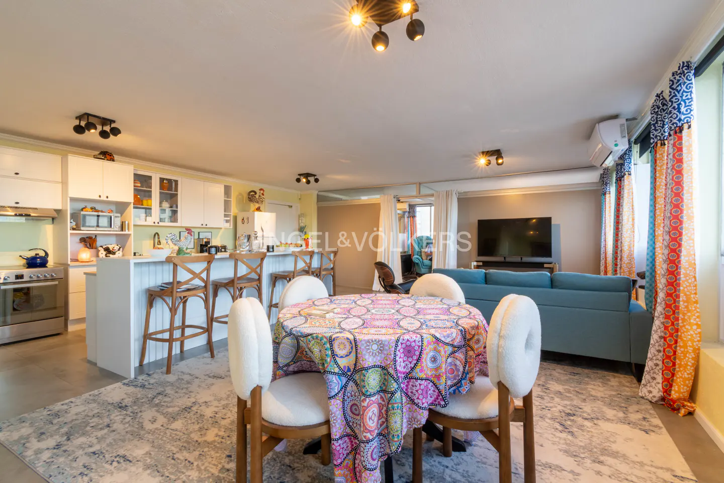 Bright, open-concept living space with a round dining table, white chairs, and a colorful tablecloth. Kitchen island, blue sofa, and TV in the background.