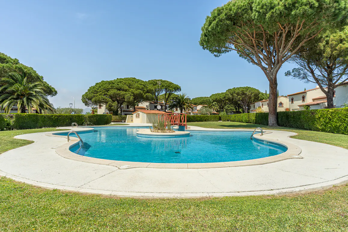 Outdoor pool with turquoise water, surrounded by green grass, trees, and hedges. Buildings with orange roofs are in the background.