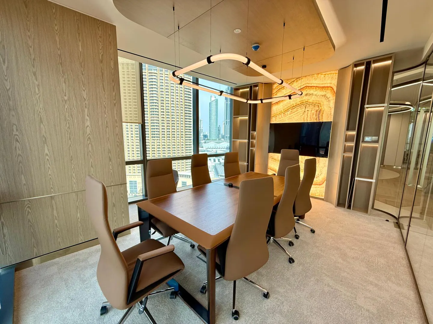 Conference room with a wood table, tan chairs, and a modern light fixture. A large window shows a city skyline.