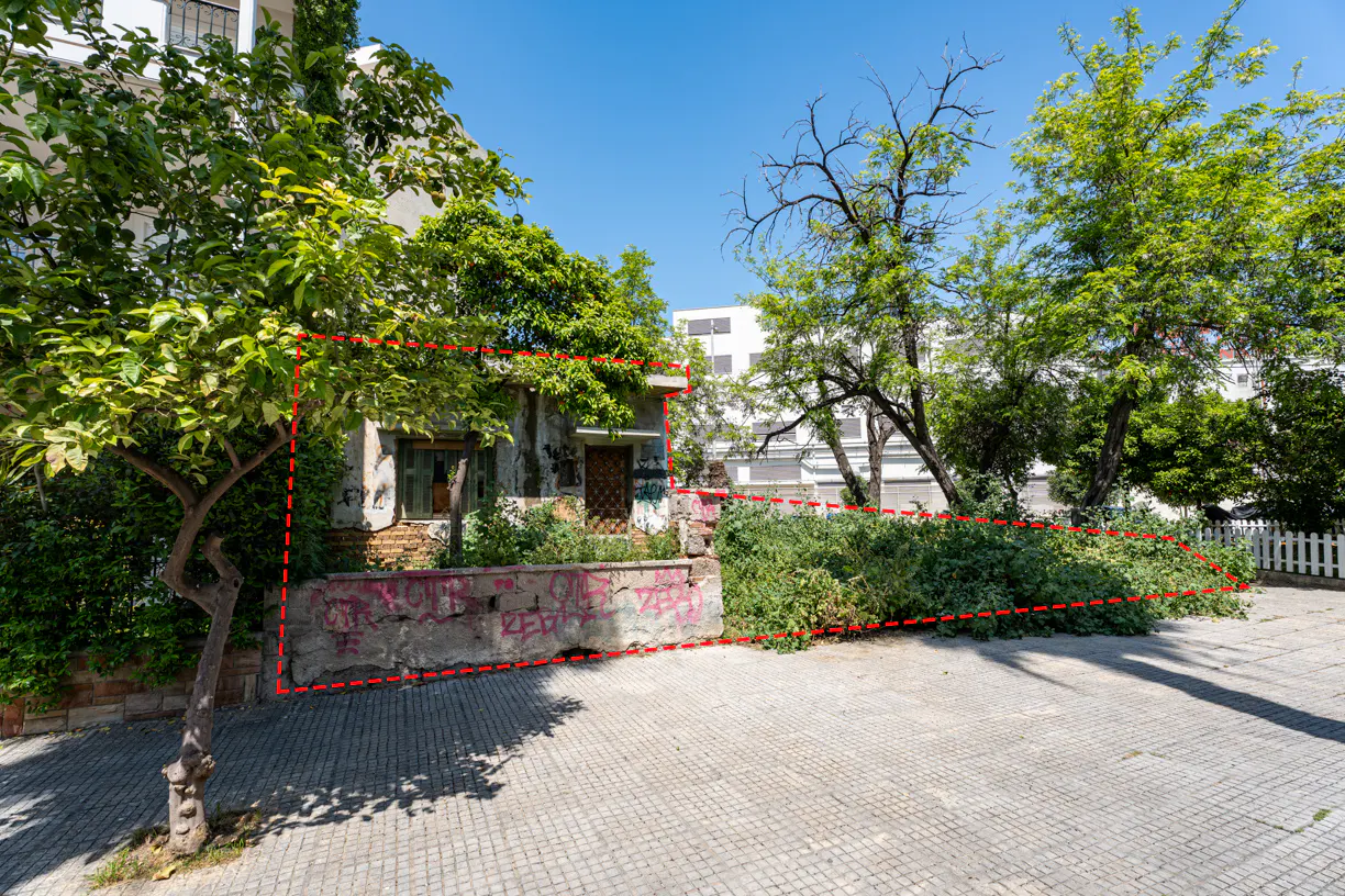 Exterior view of a small, rundown building with graffiti on the wall, surrounded by overgrown greenery and trees.