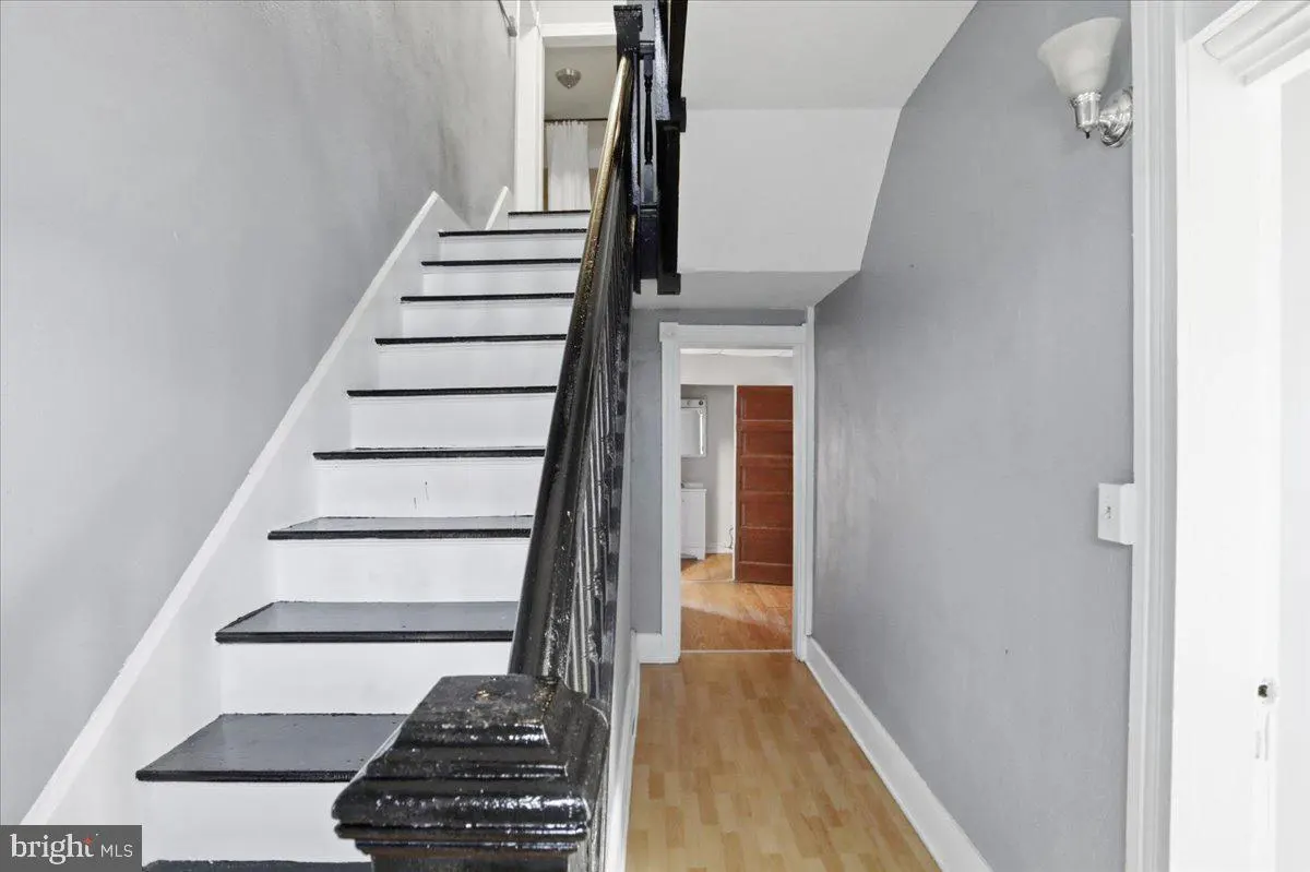 Interior view of a home's entryway with white and black stairs, a dark wood banister, and a hallway with light wood floors. Gray walls and white trim.