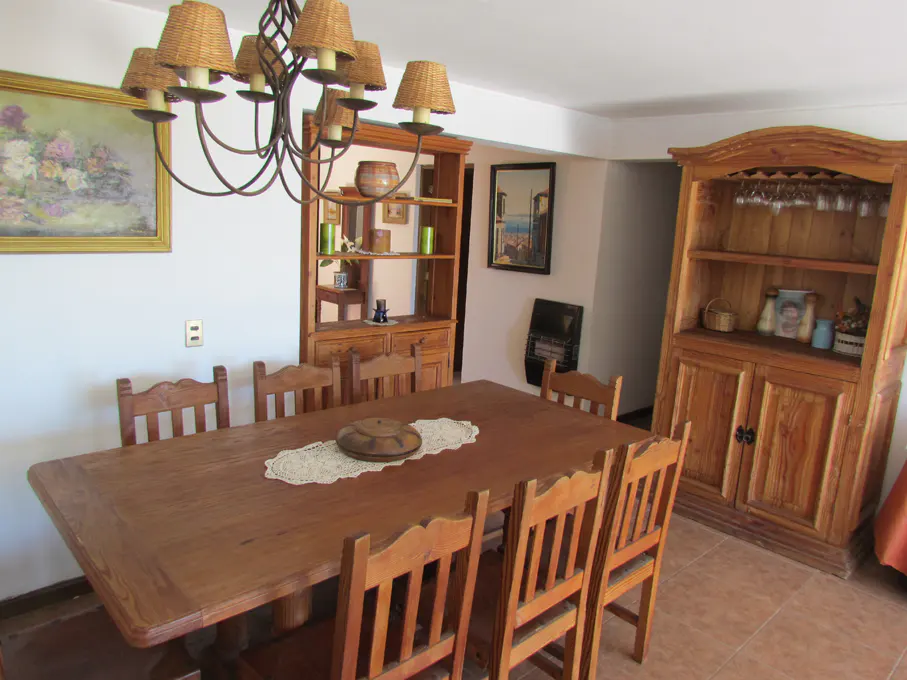 A dining room with a long wooden table, chairs, and a chandelier. Two wooden cabinets are in the background. A painting hangs on the wall.